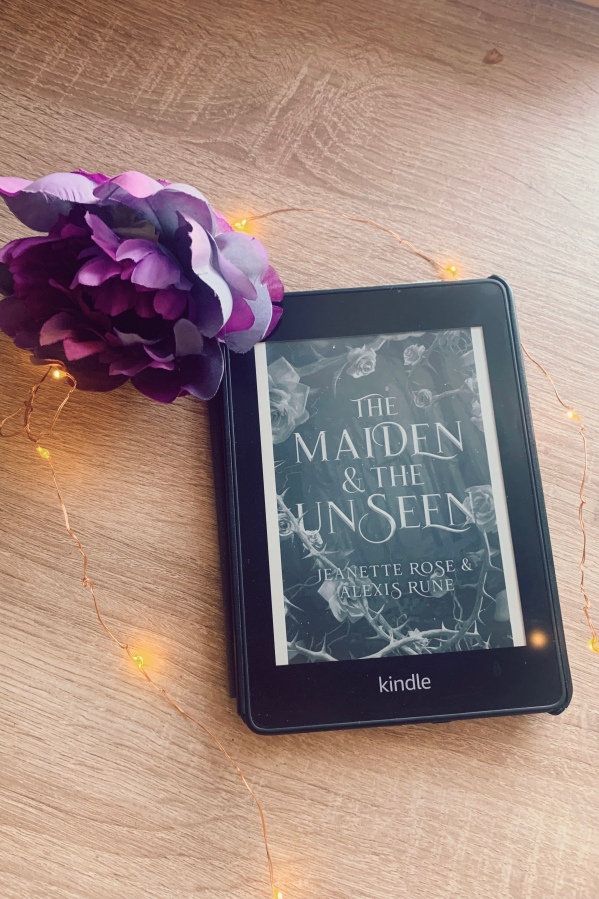 An Amazon Kindle Paperwhite showing the cover for The Maiden and the Unseen in black and white with a fake purple rose next to it and surrounded by twinkle lights on a light wood background.