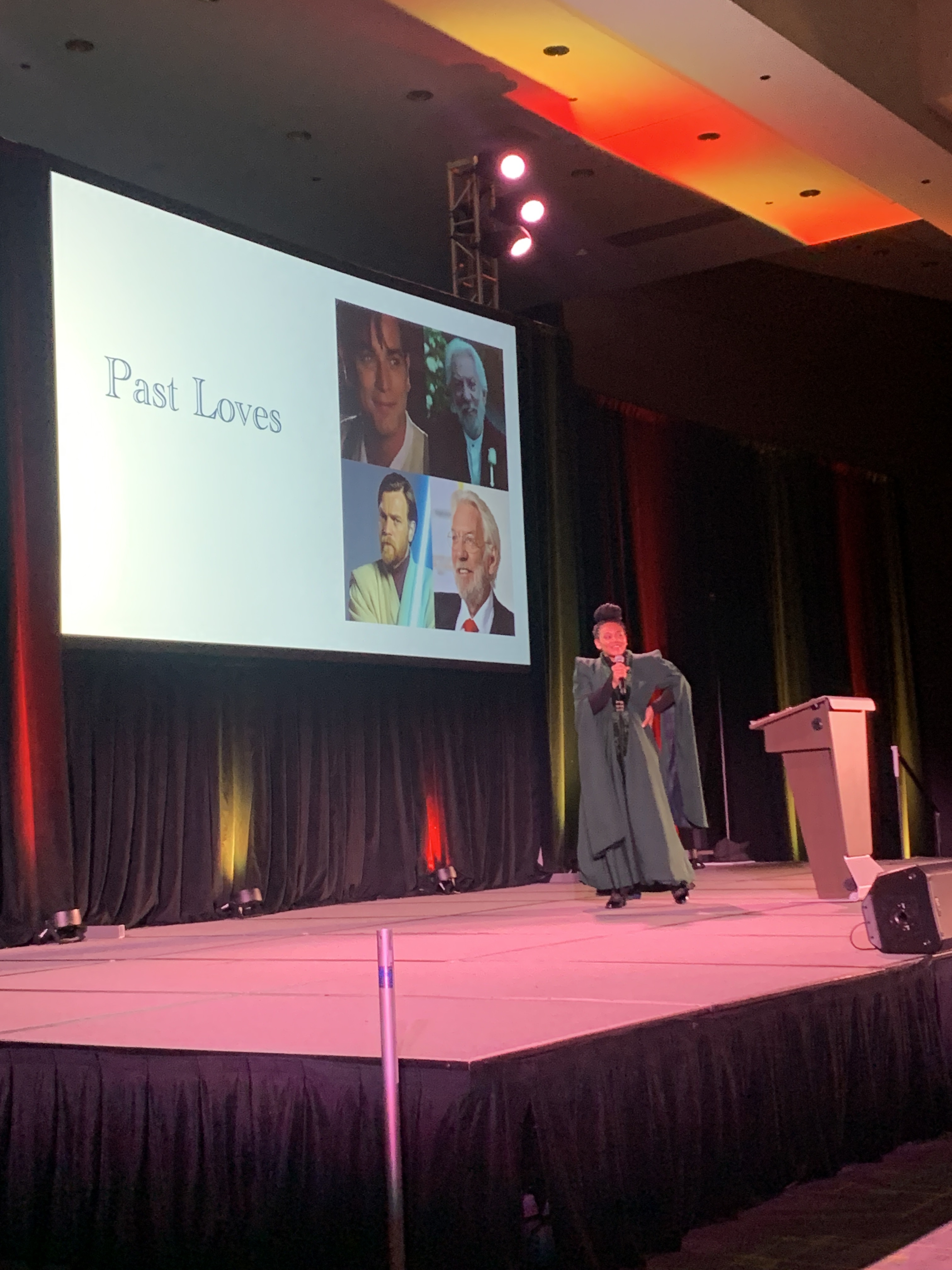 Chanel Williams dressed as Minerva McGonnagall on stage for her LeakCon Panel in front of a powerpoint slide that says past loves with photos Ewan McGregor and President Snow from the Hunger Games movies.