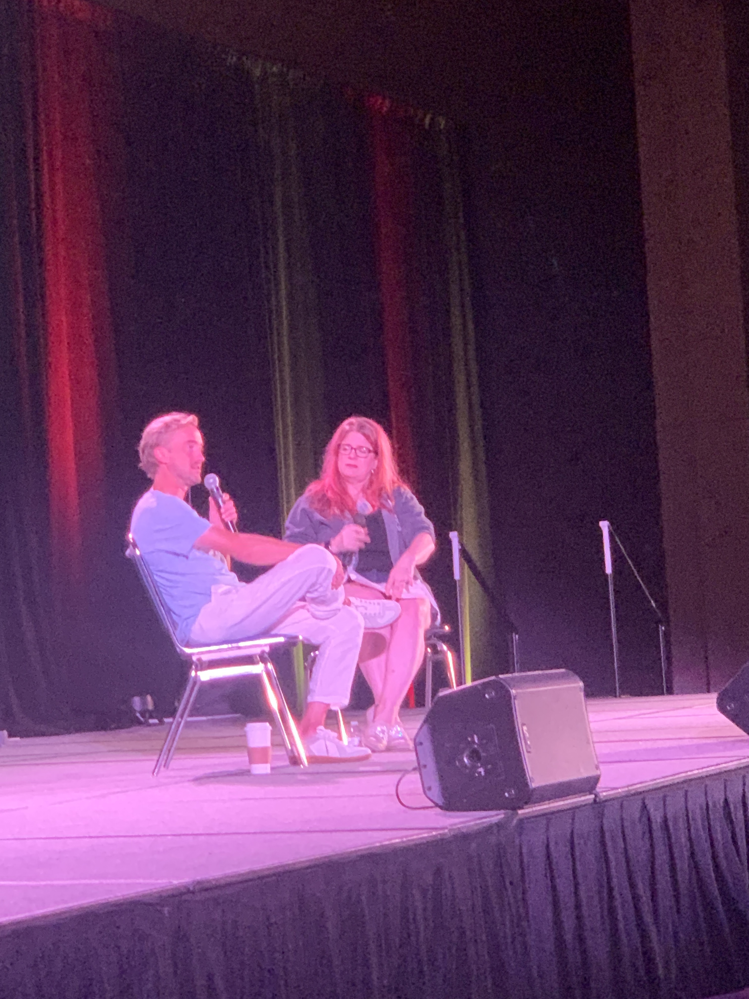 Tom Felton being interviewed on stage by Melissa Anelli, a pinkish light tints the photo.