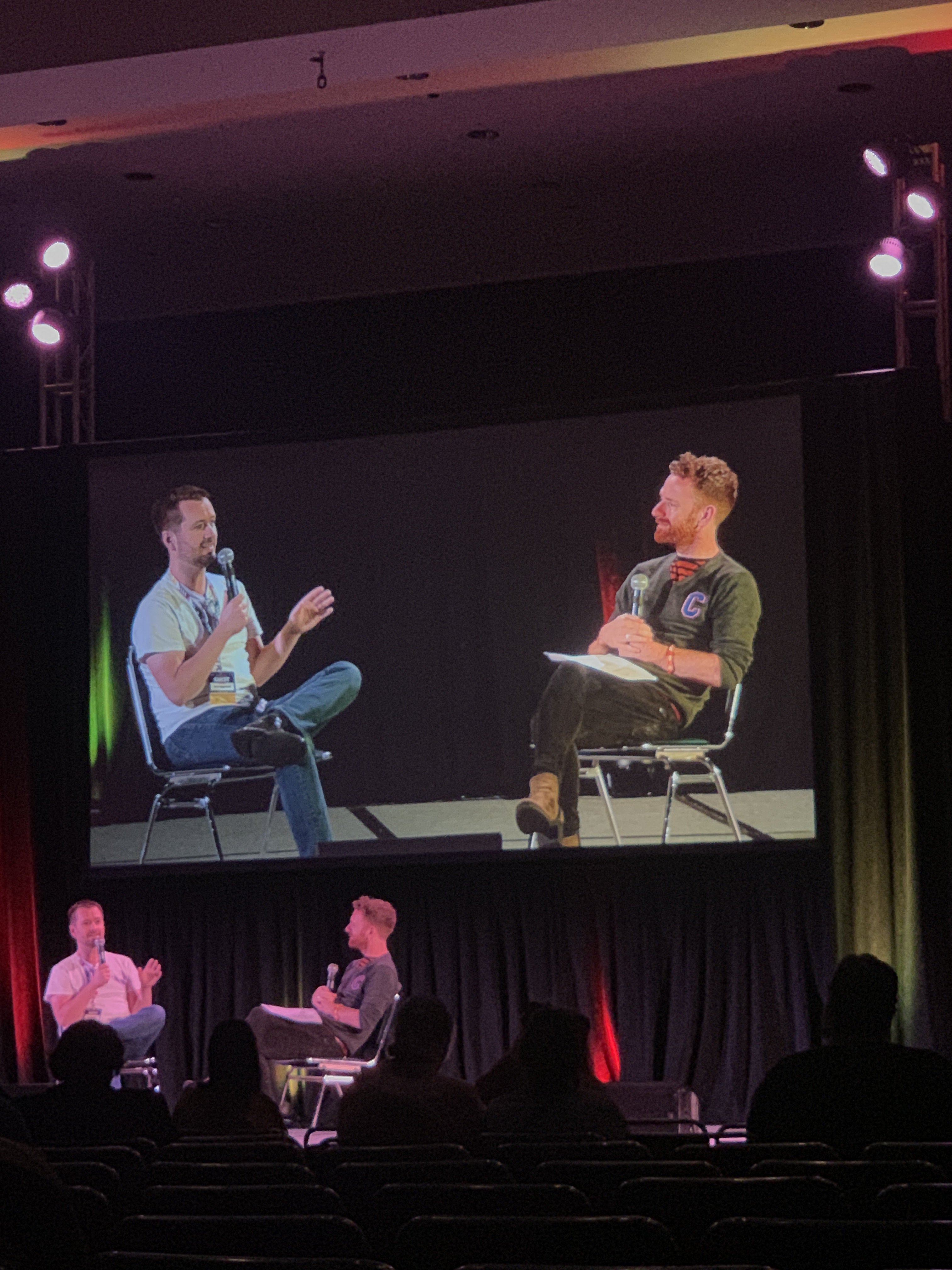 Sean Biggerstaff and Chris Rankin chatting with eachother on a stage with a video of them behind to enlarge for the rest of the room. Several people are in seats in silhouette in front of them.